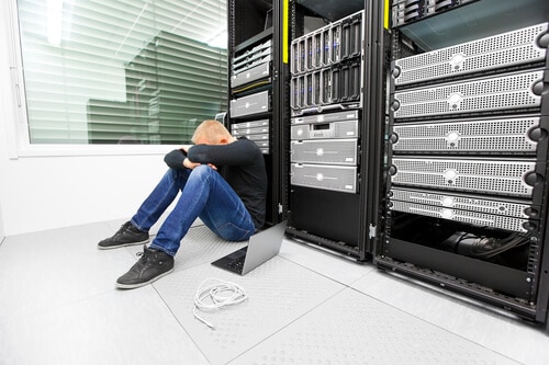 Exhausted and tired IT engineer or technician with problems sitting and leaning against data racks in a datacenter.